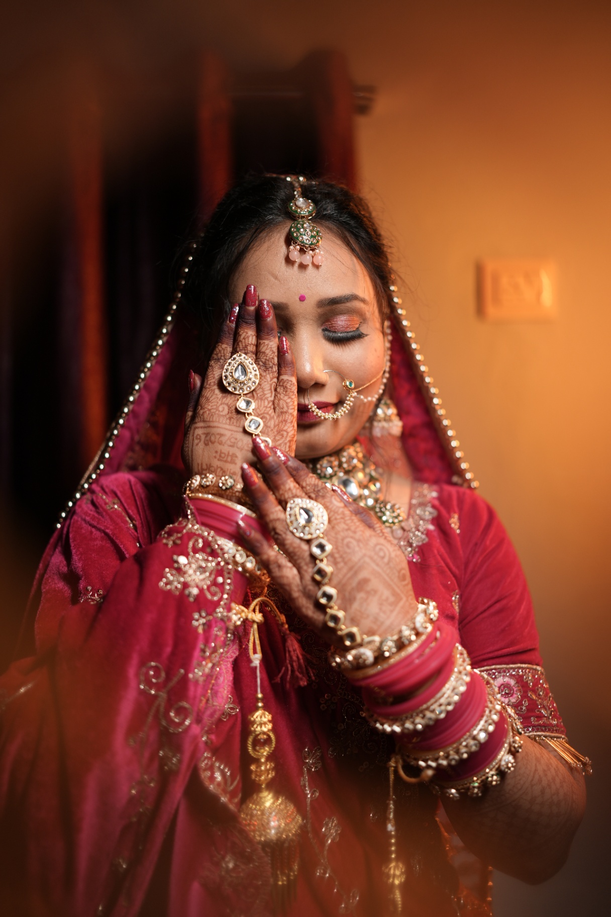 Bride resting jewelled hands against her face, mehndi and kundan in warm lamplight